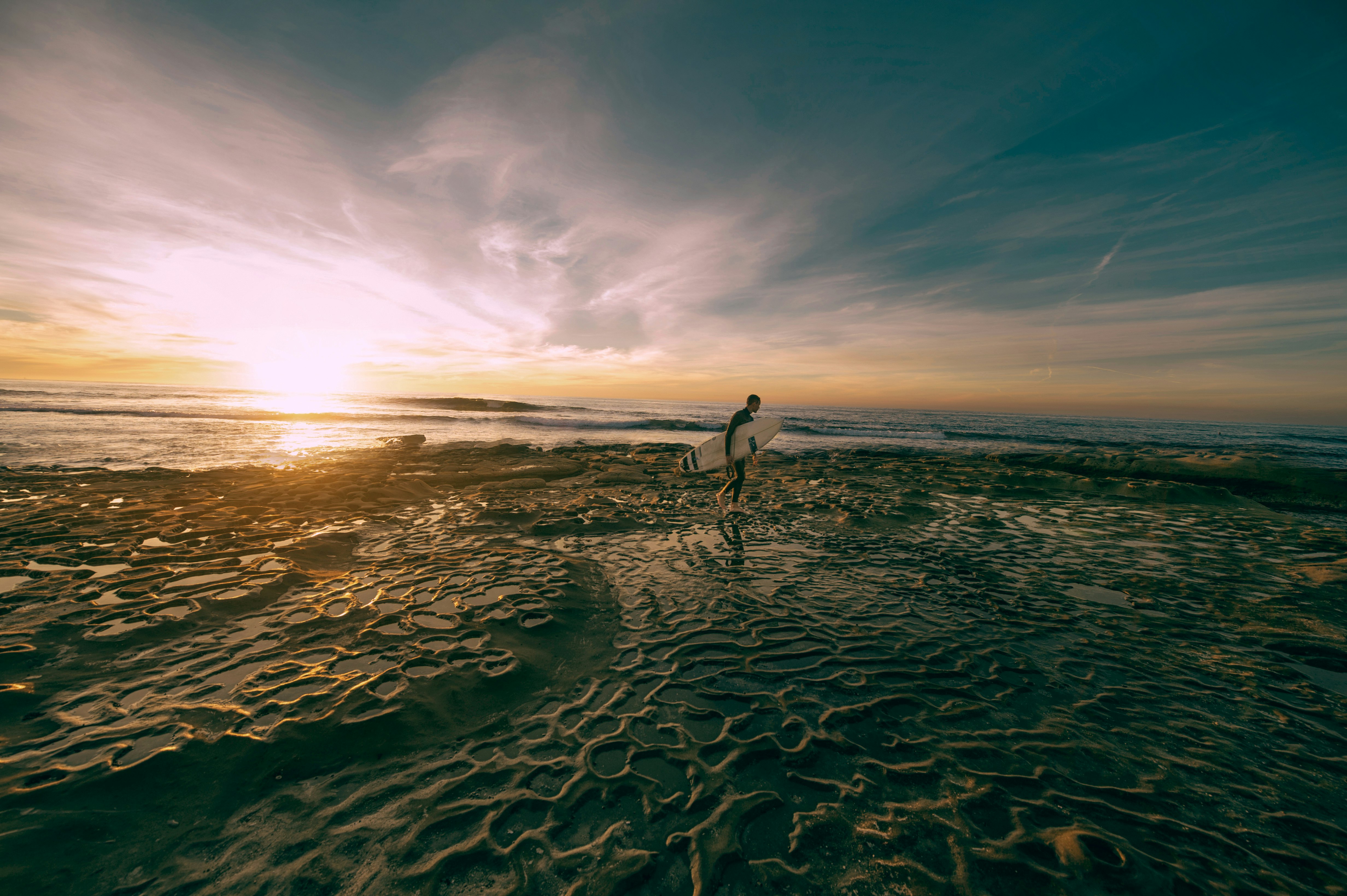 man carrying white surfboard while walking on shoreline at daytime